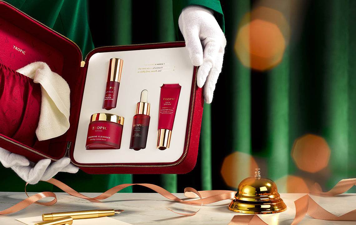 Tropic luxury skincare gift set featuring red and gold products inside a red case, held by gloved hands with festive ribbon, green backdrop, and golden bell.